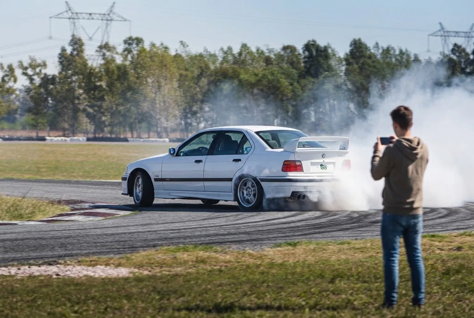 BMW E36 derrapando con humo en las gomas traseras mientras un alumno filma desde una zona segura de la pista