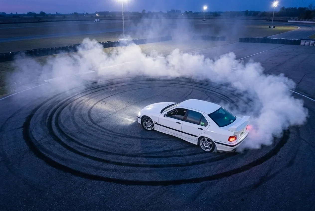 Vista elevada de un BMW E36 blanco haciendo un donut, mostrando el círculo de humo y las marcas de goma sobre el asfalto