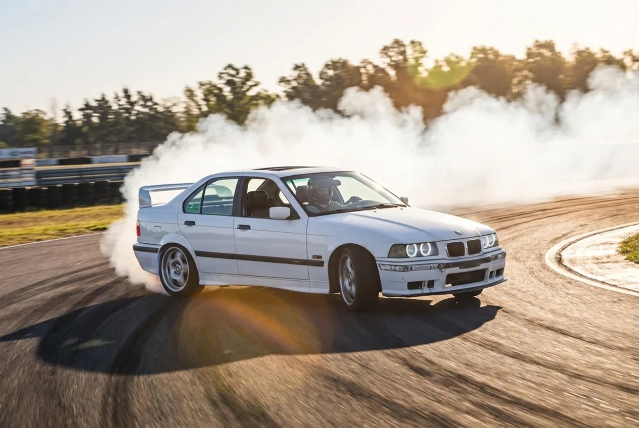 BMW E36 blanco en drift en movimiento, avanzando de costado por una curva con humo al atardecer