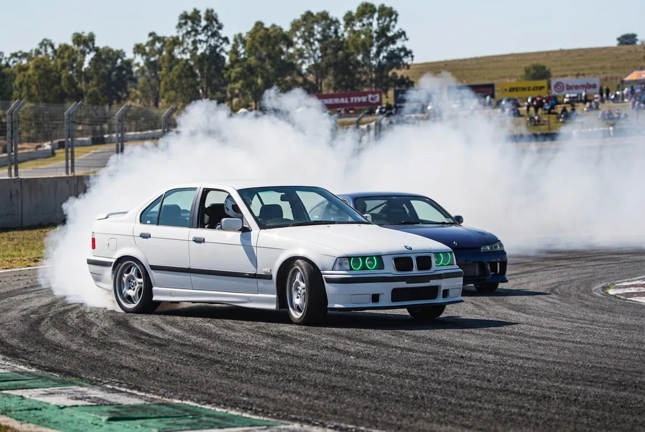 Two cars drifting in tandem inches apart, the white BMW E36 chasing through the smoke