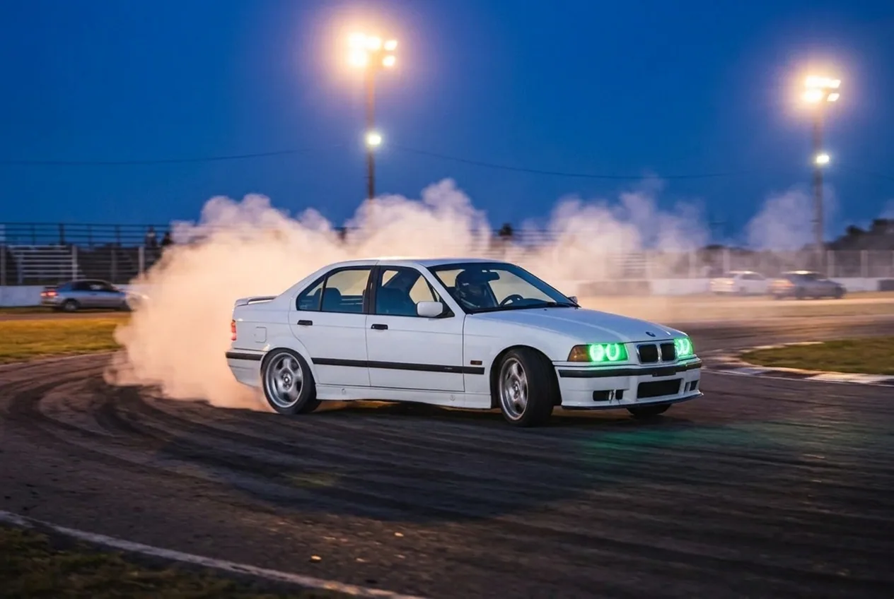BMW E36 blanco derrapando al caer la tarde bajo los reflectores del autodromo, sumando vueltas