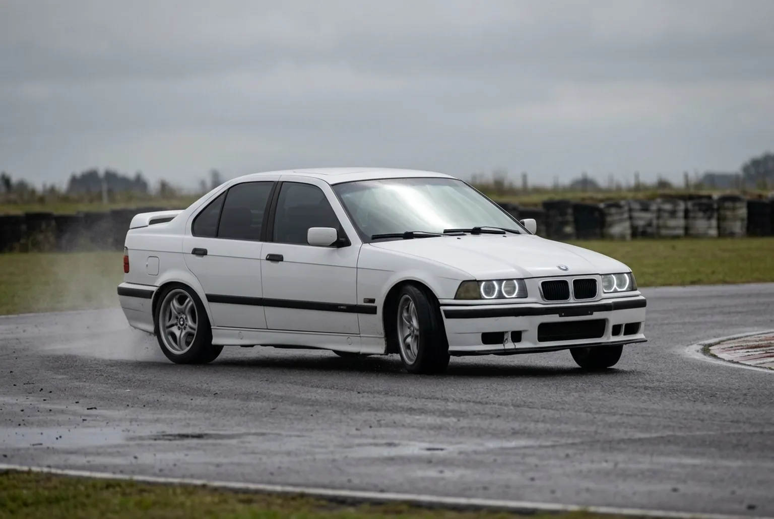 BMW E36 blanco deslizando suave sobre asfalto humedo de pista bajo un cielo gris nublado