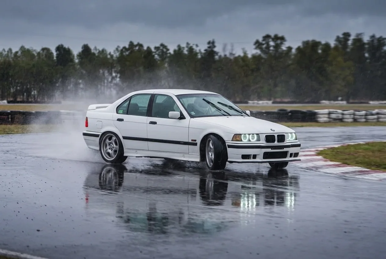 BMW E36 blanco derrapando sobre asfalto mojado y reflectante bajo la lluvia en una pista cerrada