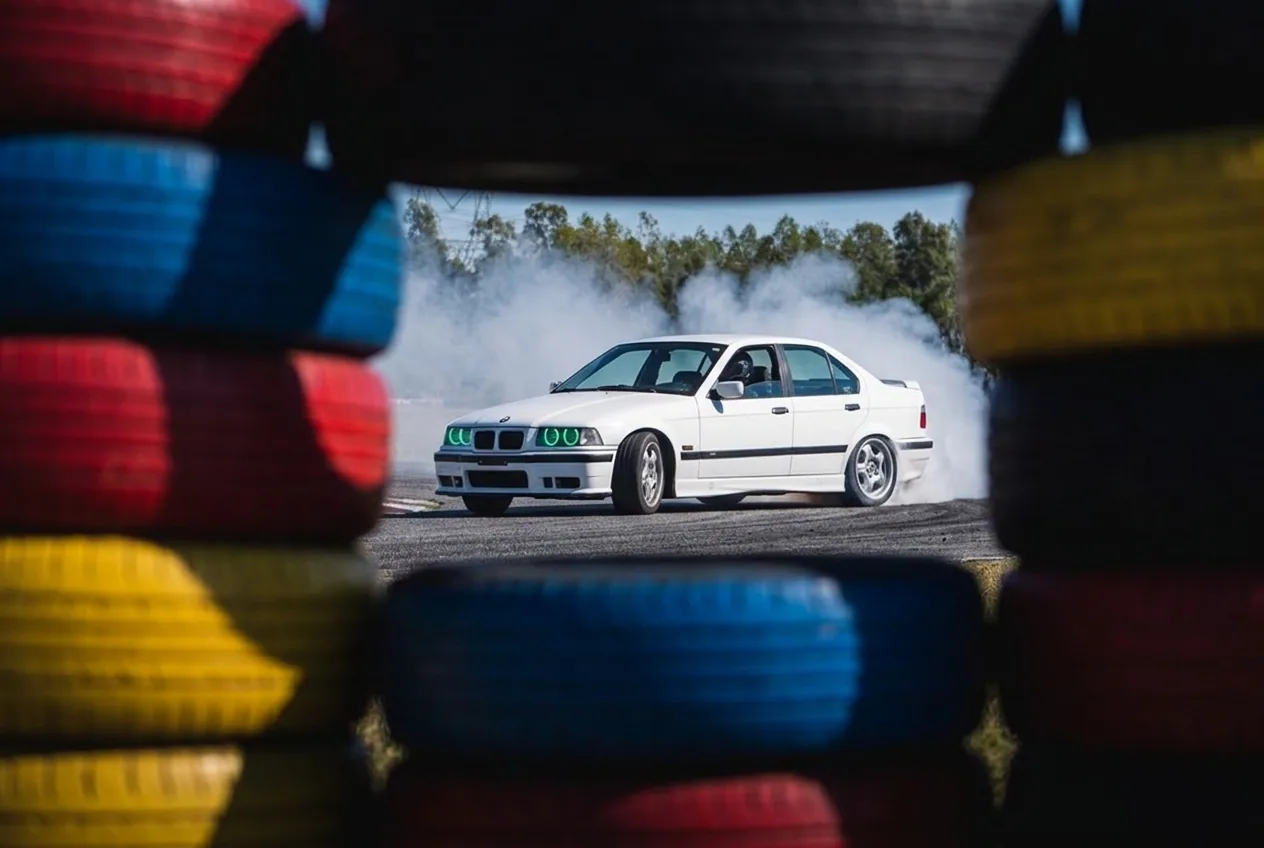 BMW E36 blanco derrapando visto a traves de una pila de cubiertas de proteccion de la pista