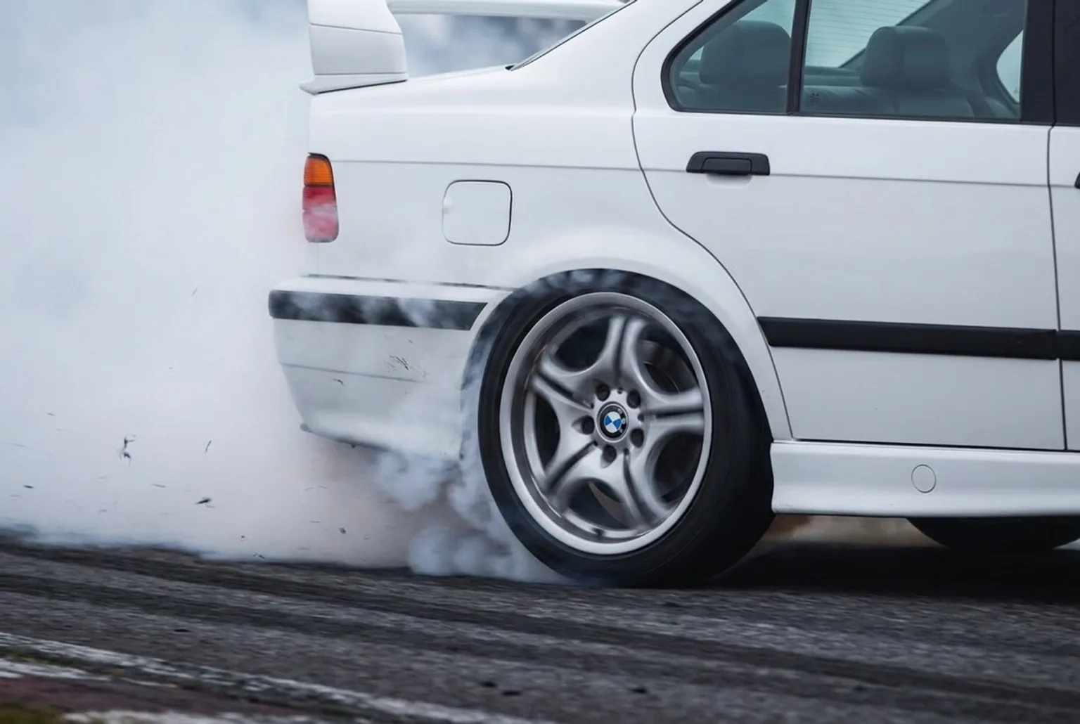 Detalle de la rueda trasera del BMW E36 blanco largando humo durante un derrape en pista cerrada