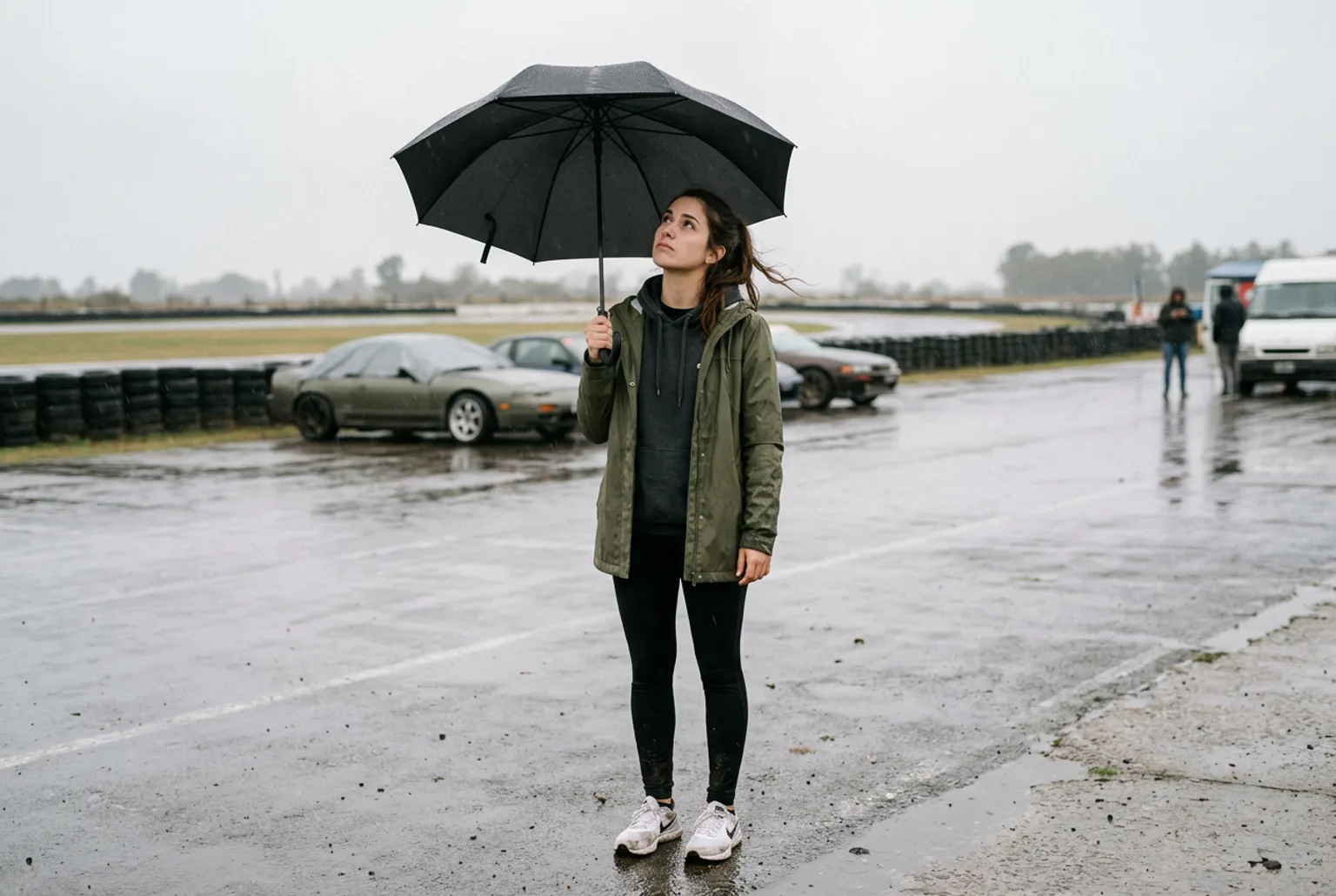 Alumna con campera impermeable verde mirando el cielo gris bajo un paraguas en el paddock mojado del autodromo