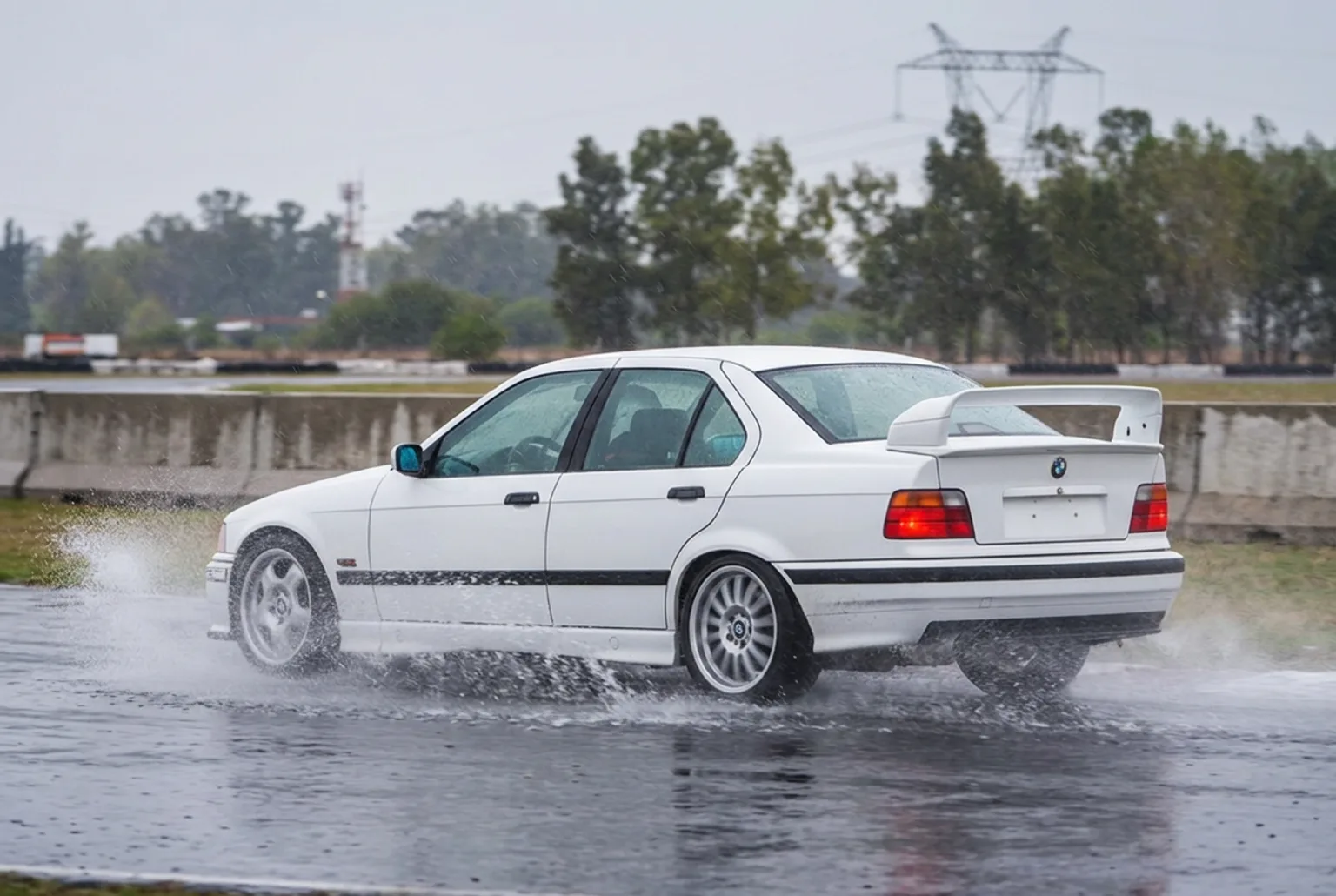 Cola de agua levantada por las ruedas traseras del BMW E36 blanco cruzado en una curva mojada del autodromo