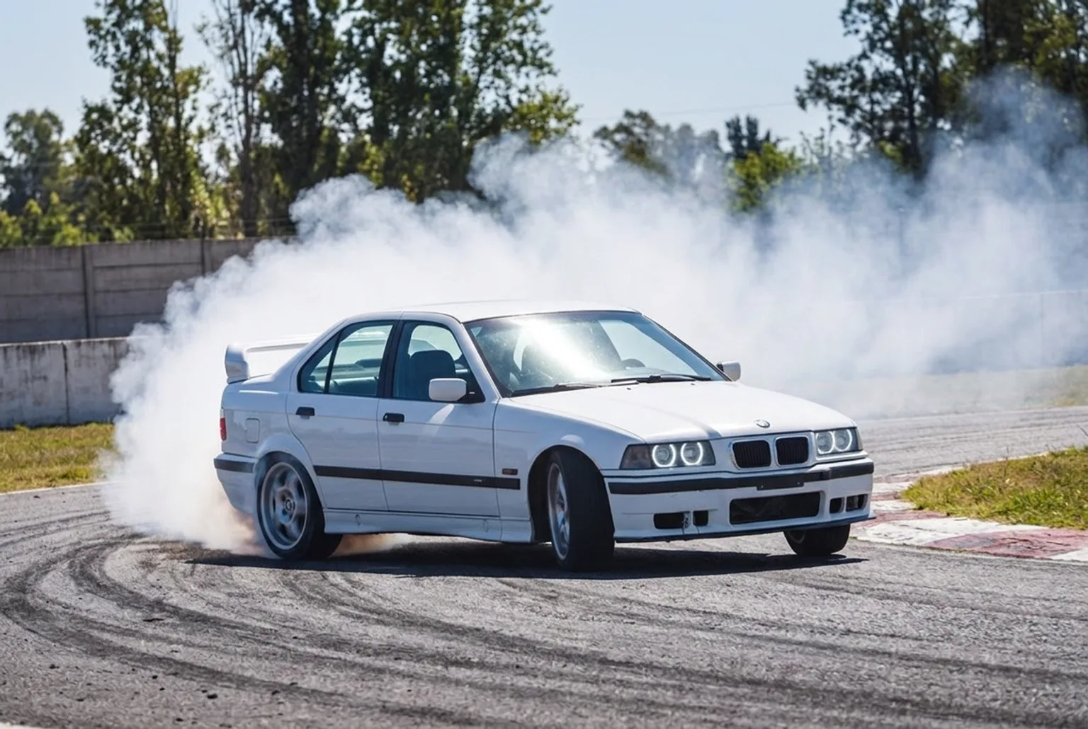 BMW E36 blanco derrapando en una curva del autodromo con humo denso saliendo de las gomas traseras, visto desde arriba en un dia soleado