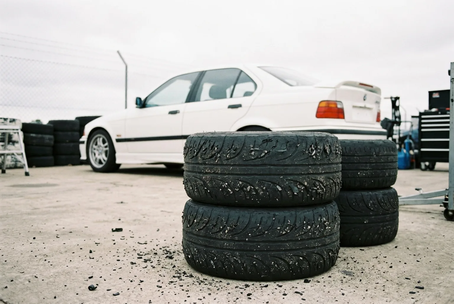 Pila de gomas de drift gastadas con el caucho destrozado en primer plano y el BMW E36 blanco desenfocado al fondo en el paddock