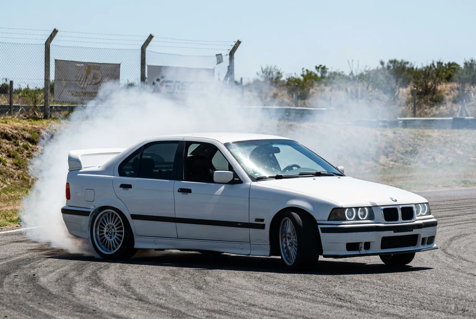 BMW E36 blanco de la escuela cruzado en pleno drift con contravolante y humo de las gomas traseras bajo el sol fuerte del mediodia