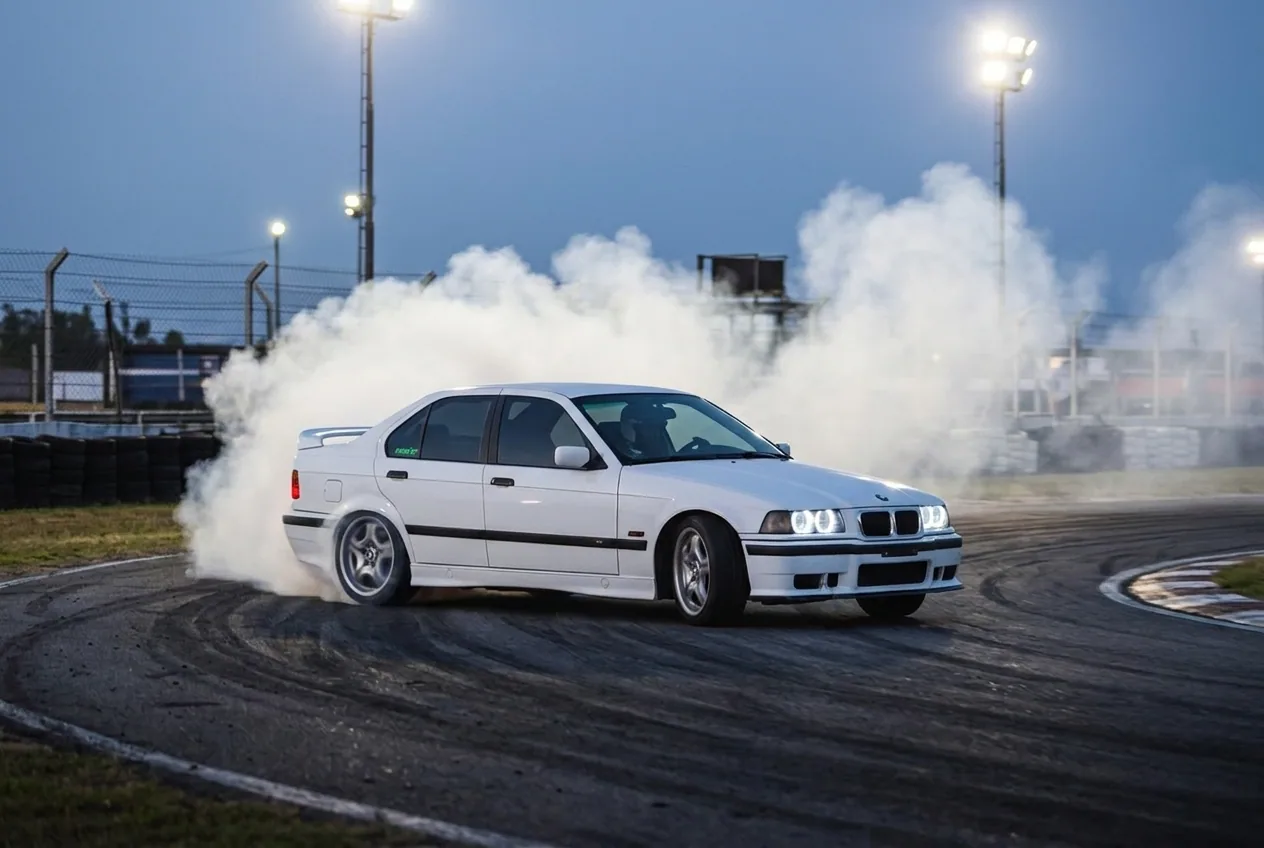 BMW E36 blanco derrapando con humo bajo las luces de la pista al anochecer, la adrenalina del regalo