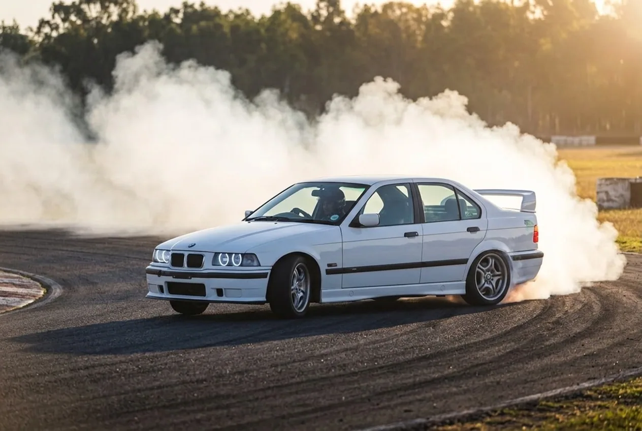 BMW E36 blanco en un drift fluido iniciado con acelerador en una curva rápida con mucho humo al atardecer