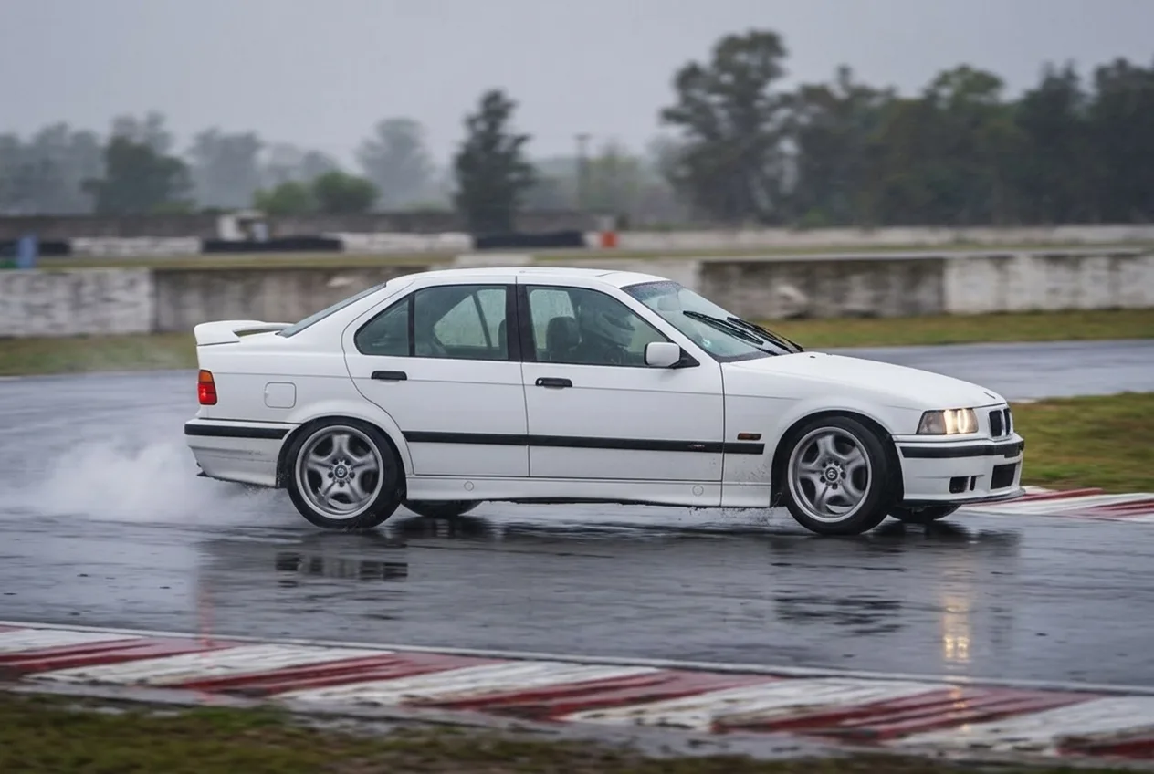 BMW E36 blanco de costado iniciando un derrape, la cola saliendo con humo en las ruedas traseras, sobre asfalto mojado en una pista cerrada de Buenos Aires