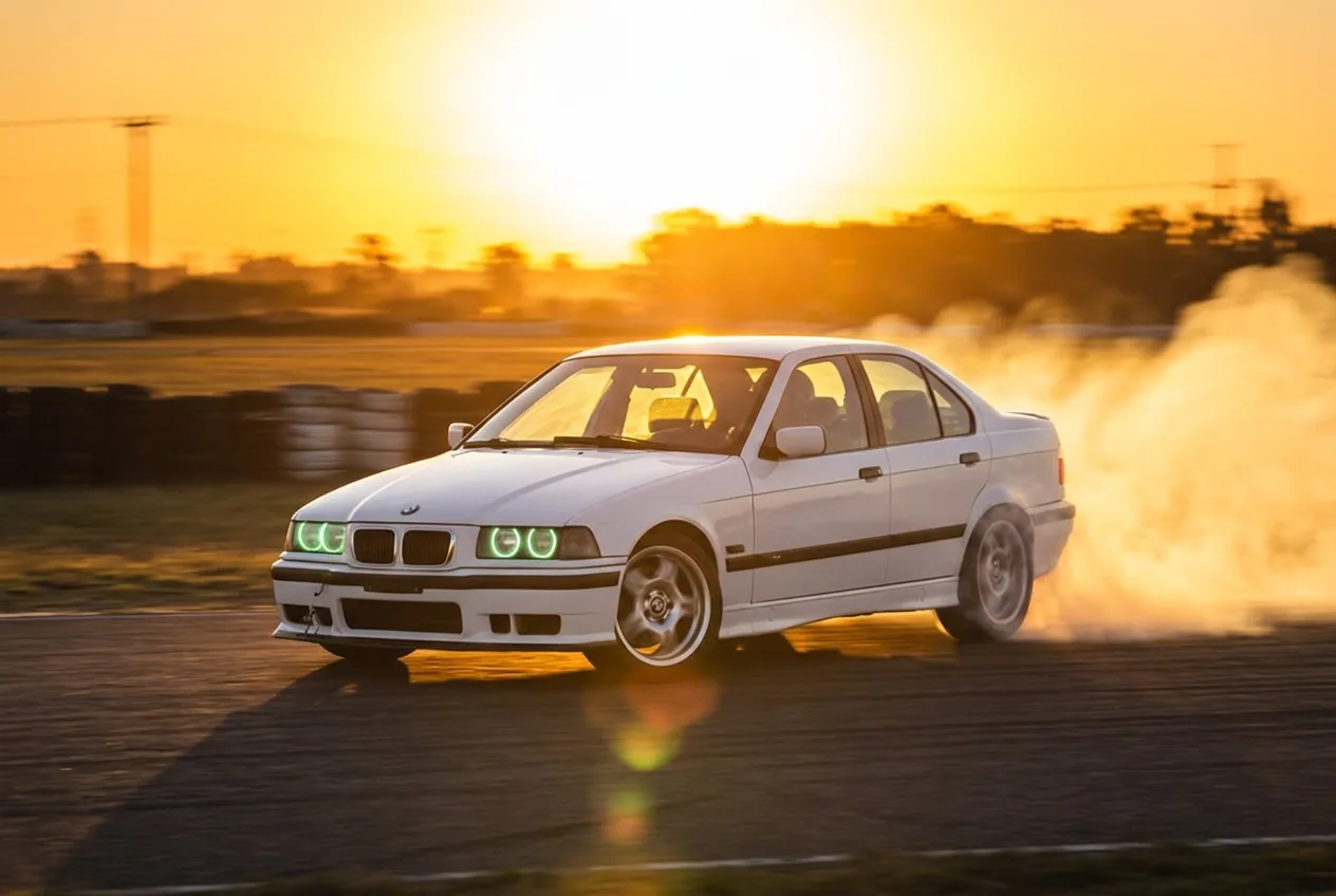 BMW E36 blanco derrapando a contraluz al atardecer con una estela de humo dorada
