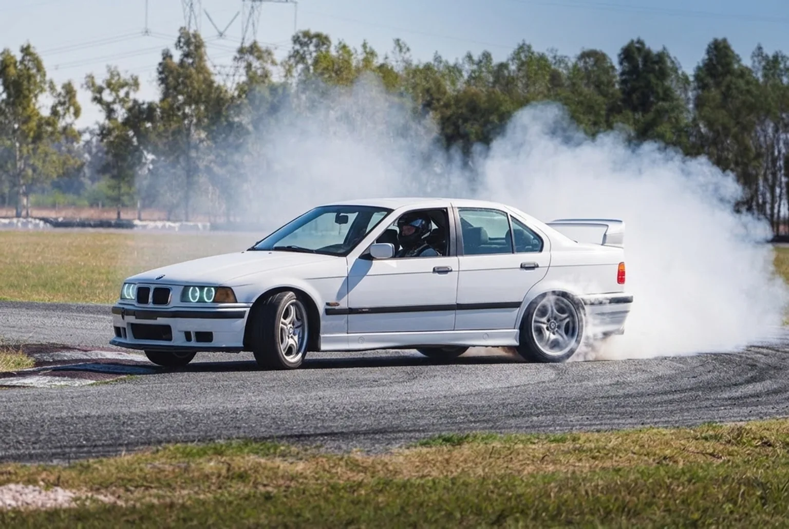 BMW E36 blanco derrapando con gran nube de humo de neumáticos bajo sol fuerte en una pista cerrada