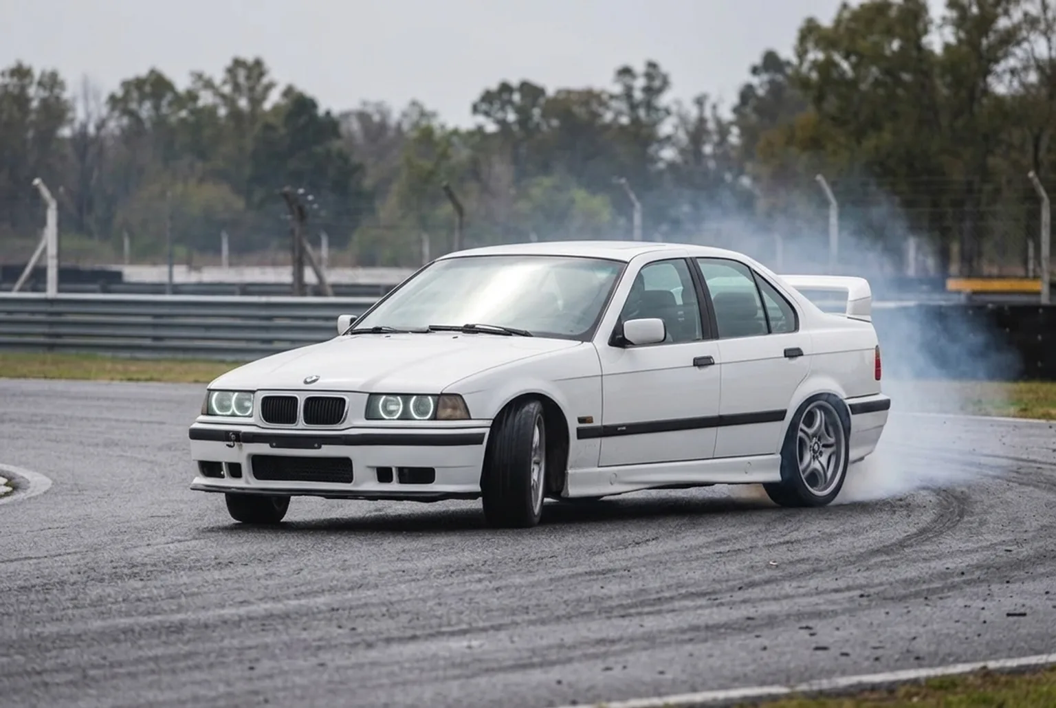 BMW E36 blanco en pleno drift con el volante contravolanteado y humo, bajo cielo gris nublado en pista cerrada