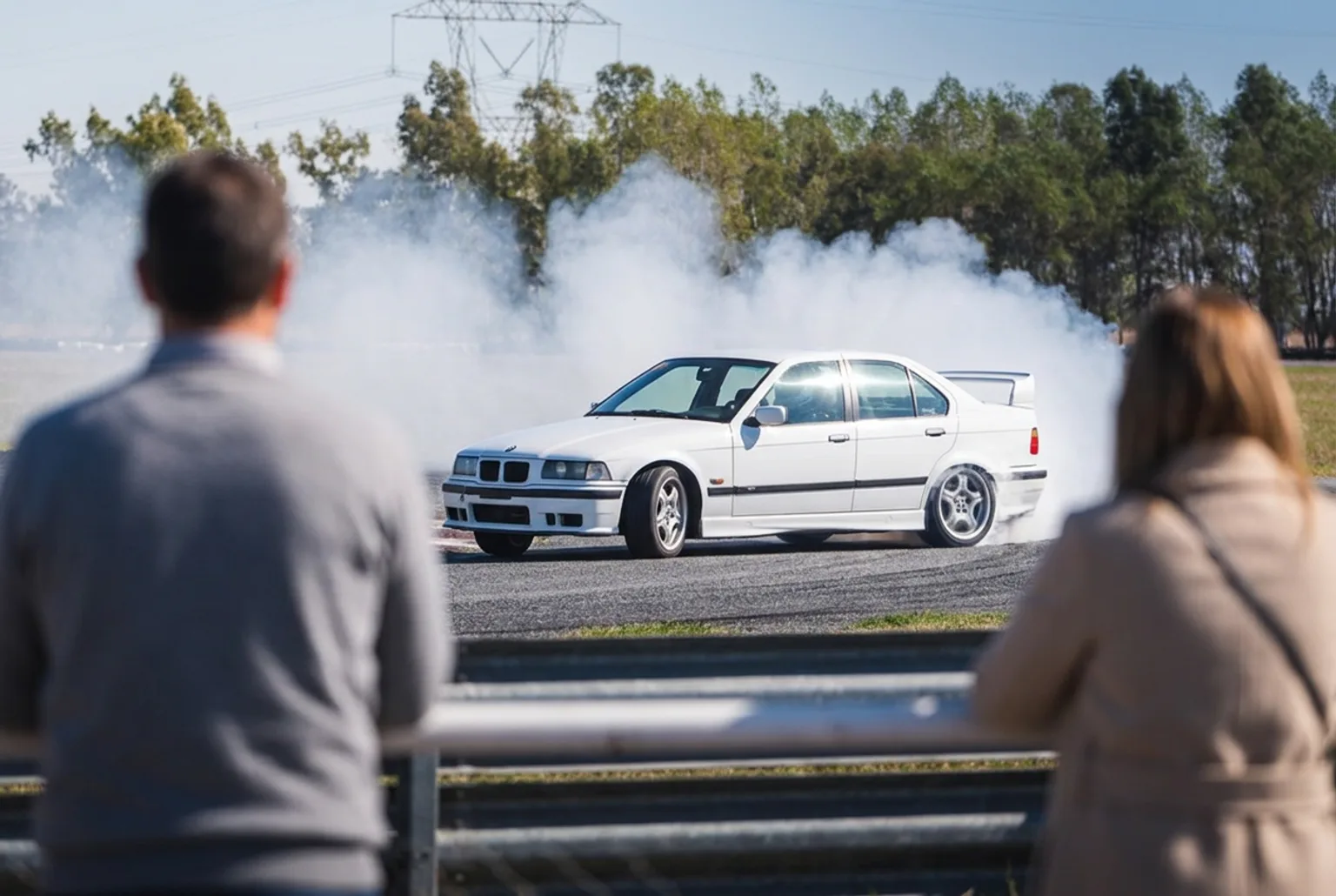 Padres desenfocados mirando desde el guardrail mientras el BMW E36 blanco derrapa en la pista con humo blanco al fondo, en un dia soleado