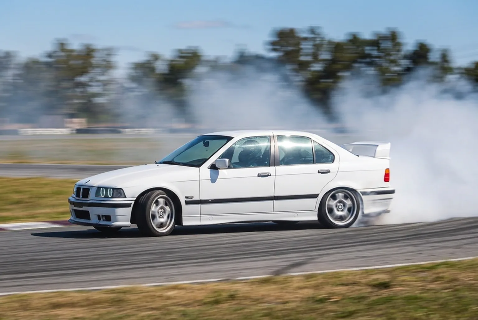 BMW E36 blanco pasando de costado en pleno derrape con estela de humo blanco, panorama con fondo movido y cielo azul de mediodia