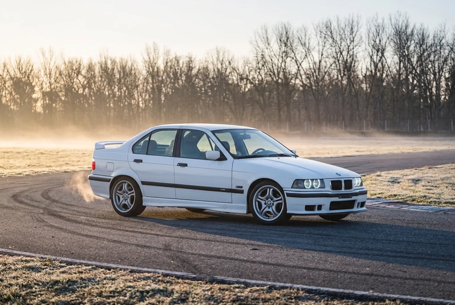 BMW E36 blanco estacionado en el asfalto frio de un autodromo al amanecer de invierno, con neblina baja y escarcha en el pasto
